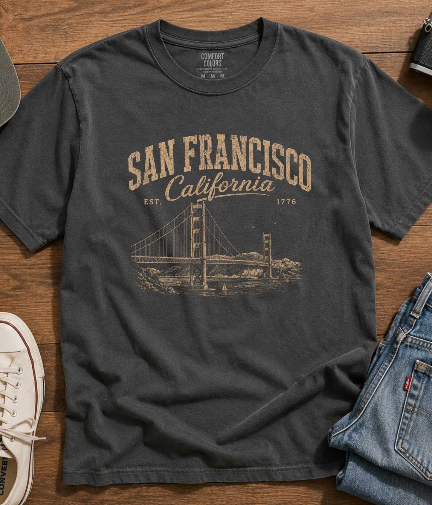 Vintage washed charcoal San Francisco California graphic t-shirt with Golden Gate Bridge design styled with denim jeans and sneakers on a wood background.