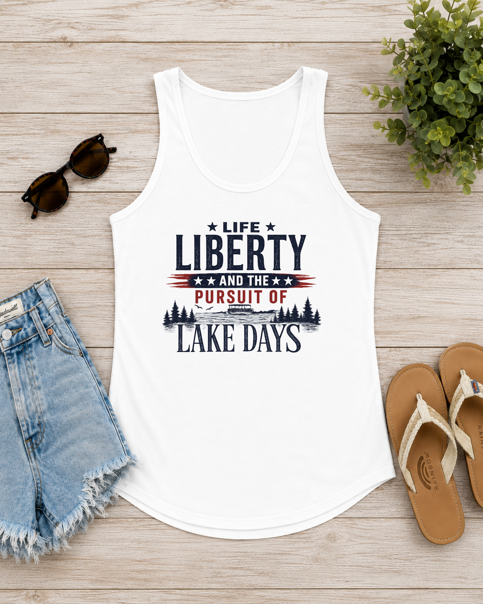 White tank top with 'Life Liberty and the Pursuit of Lake Days' text on a wooden surface with sunglasses, shorts, and sandals.