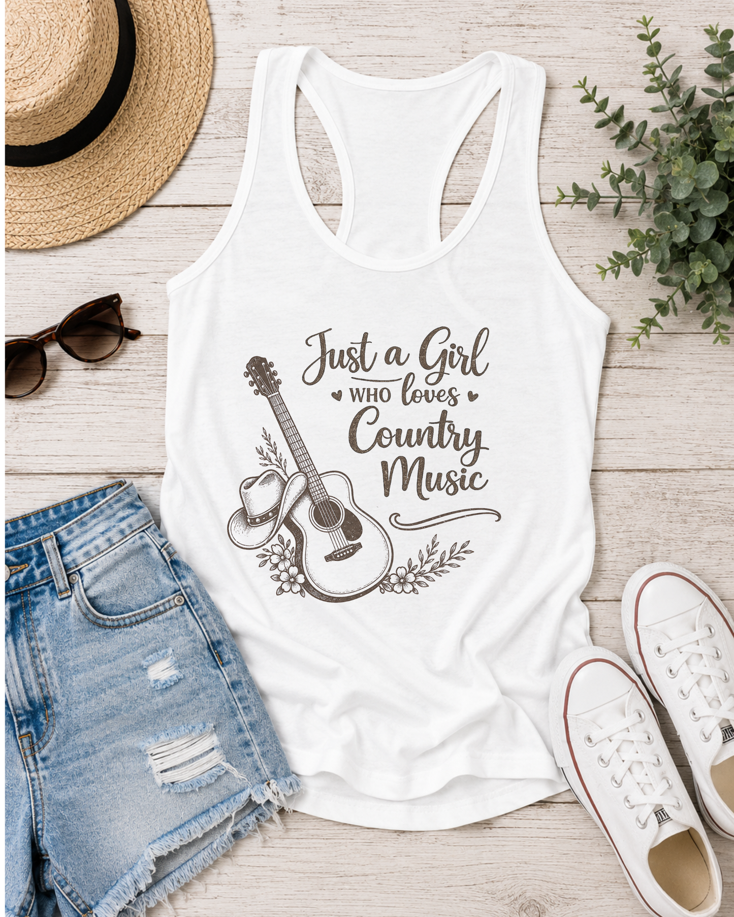 White racerback tank top with 'Just a Girl Who Loves Country Music' text and graphics on a wooden surface with shorts, sneakers, and a hat.