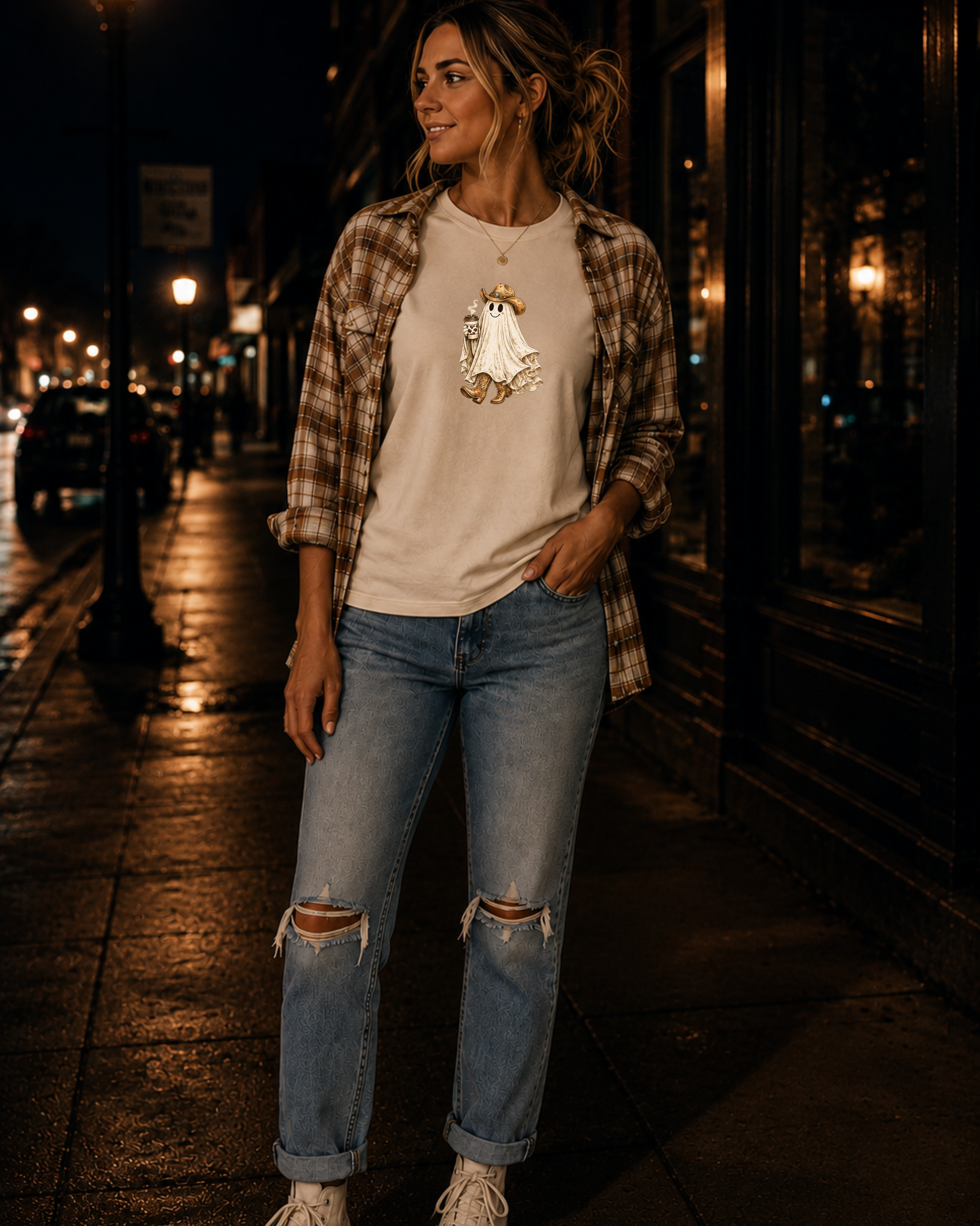 Woman standing on the sidewalk at night wearing a vintage western ghost graphic on a natural t-shirt featuring a smiling ghost in a cowboy hat and cowboy boots holding a cow print coffee cup.