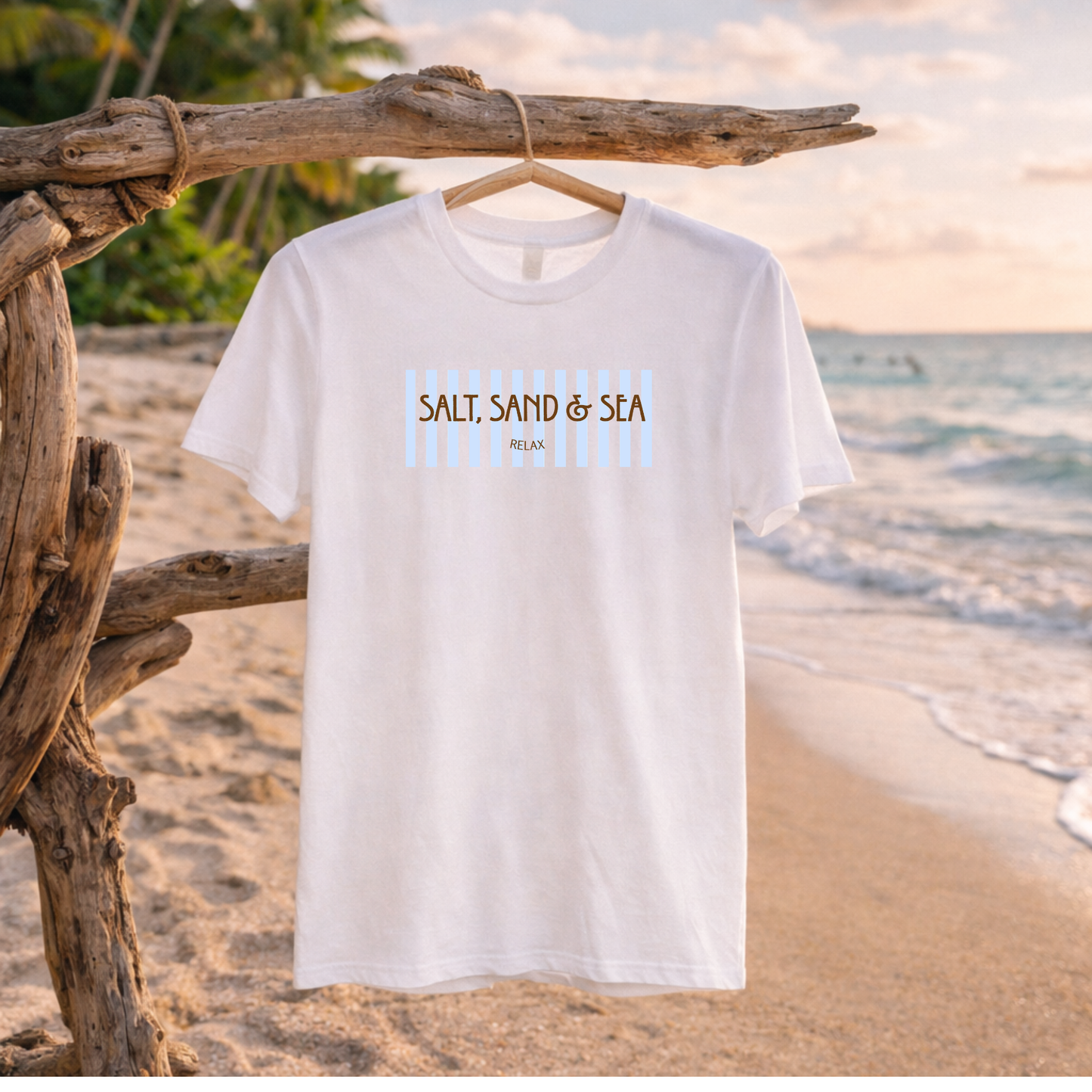 A white short-sleeve t-shirt with a 'SALT SAND & SEA' graphic hanging on a wooden branch at a beach. The graphic features brown text and light blue vertical stripes.