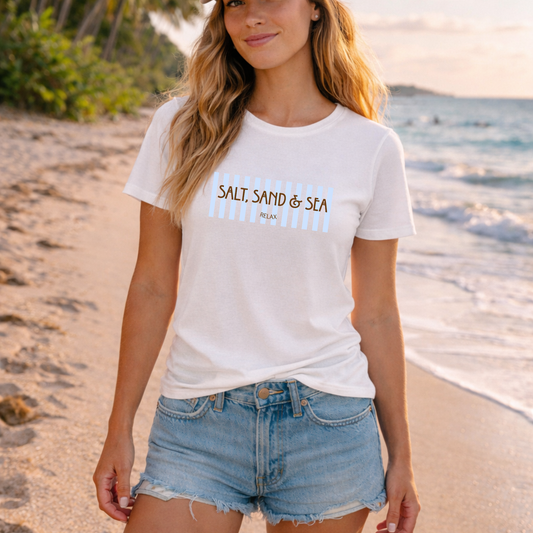 A woman wearing a white graphic t-shirt with 'SALT SAND & SEA' printed on the chest, paired with denim shorts, standing on a sandy beach with the ocean in the background.