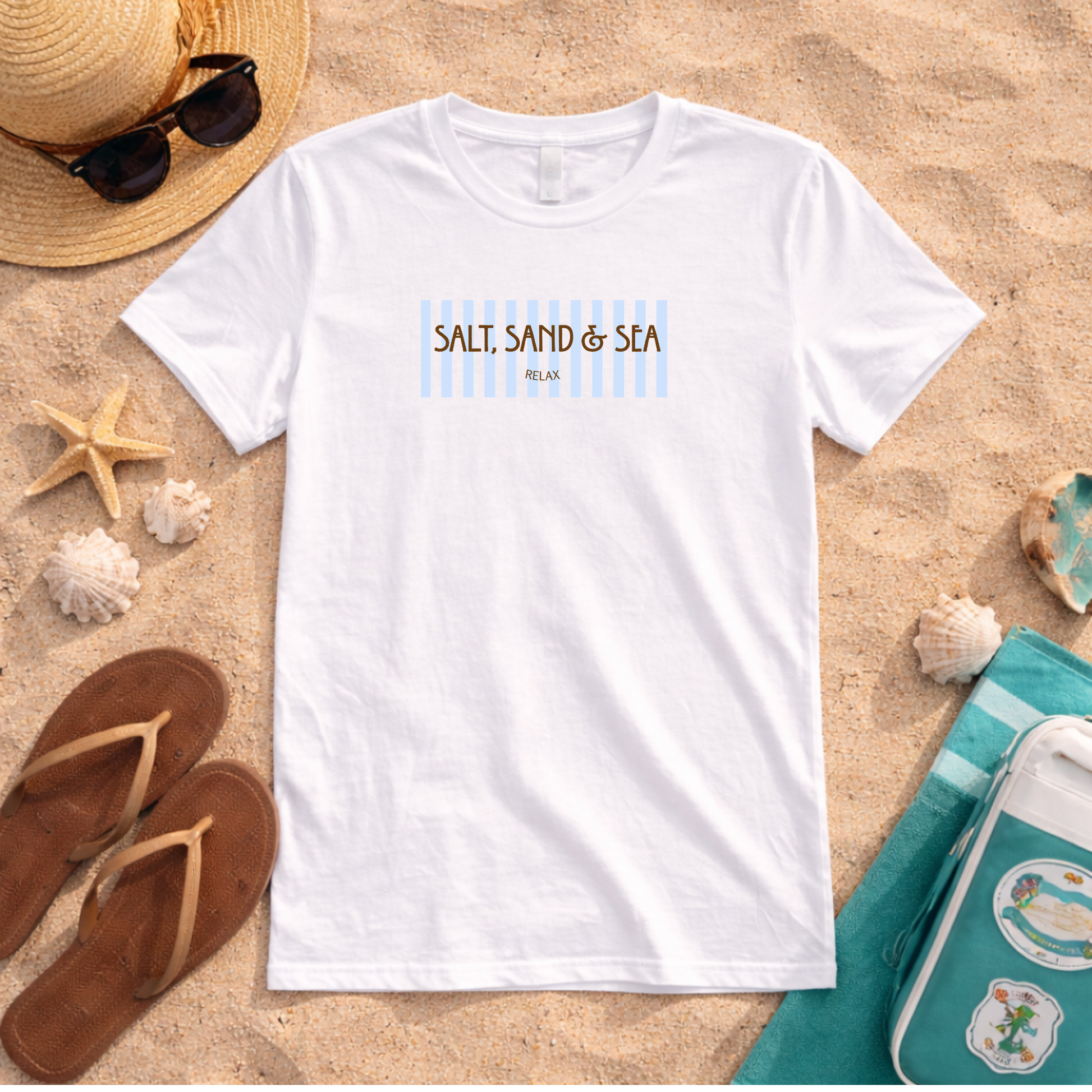A white short-sleeve graphic T-shirt laid flat on sand, surrounded by beach-themed props including a straw hat with sunglasses, flip-flops, shells, and a turquoise towel. The shirt has a chest graphic reading “SALT, SAND & SEA” with light blue vertical stripes and small brown text below.