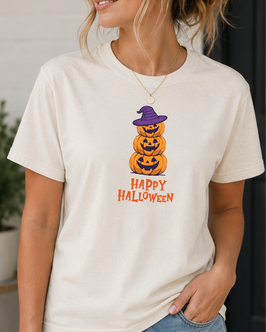 A woman wearing a cream-colored short-sleeve t-shirt with a graphic of three stacked jack-o'-lanterns wearing a purple witch hat and the text 'HAPPY HALLOWEEN' printed below in orange.