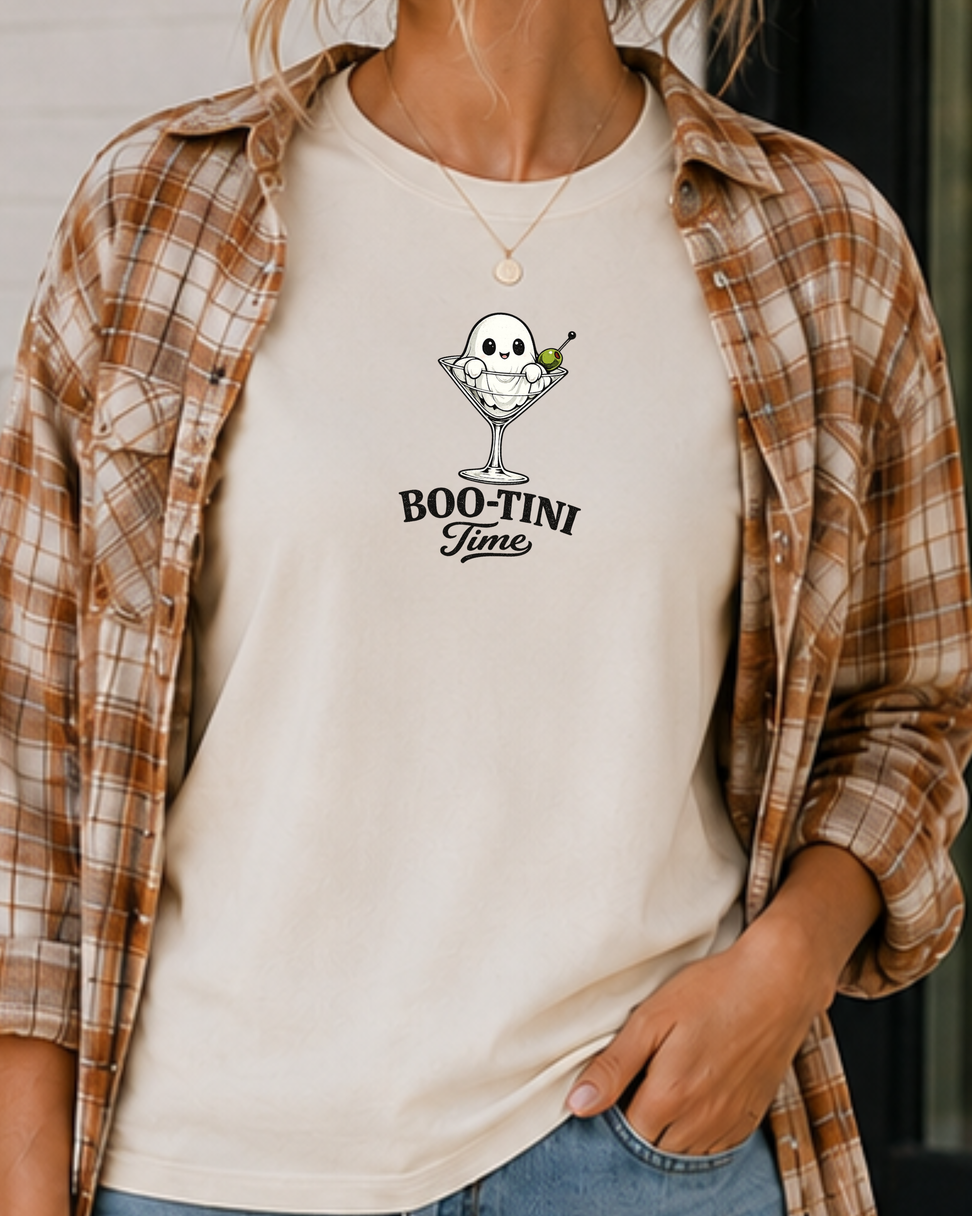 A woman wearing a cream-colored t-shirt with a 'BOO-TINI Time' martini ghost graphic, layered under a brown and beige plaid flannel shirt.