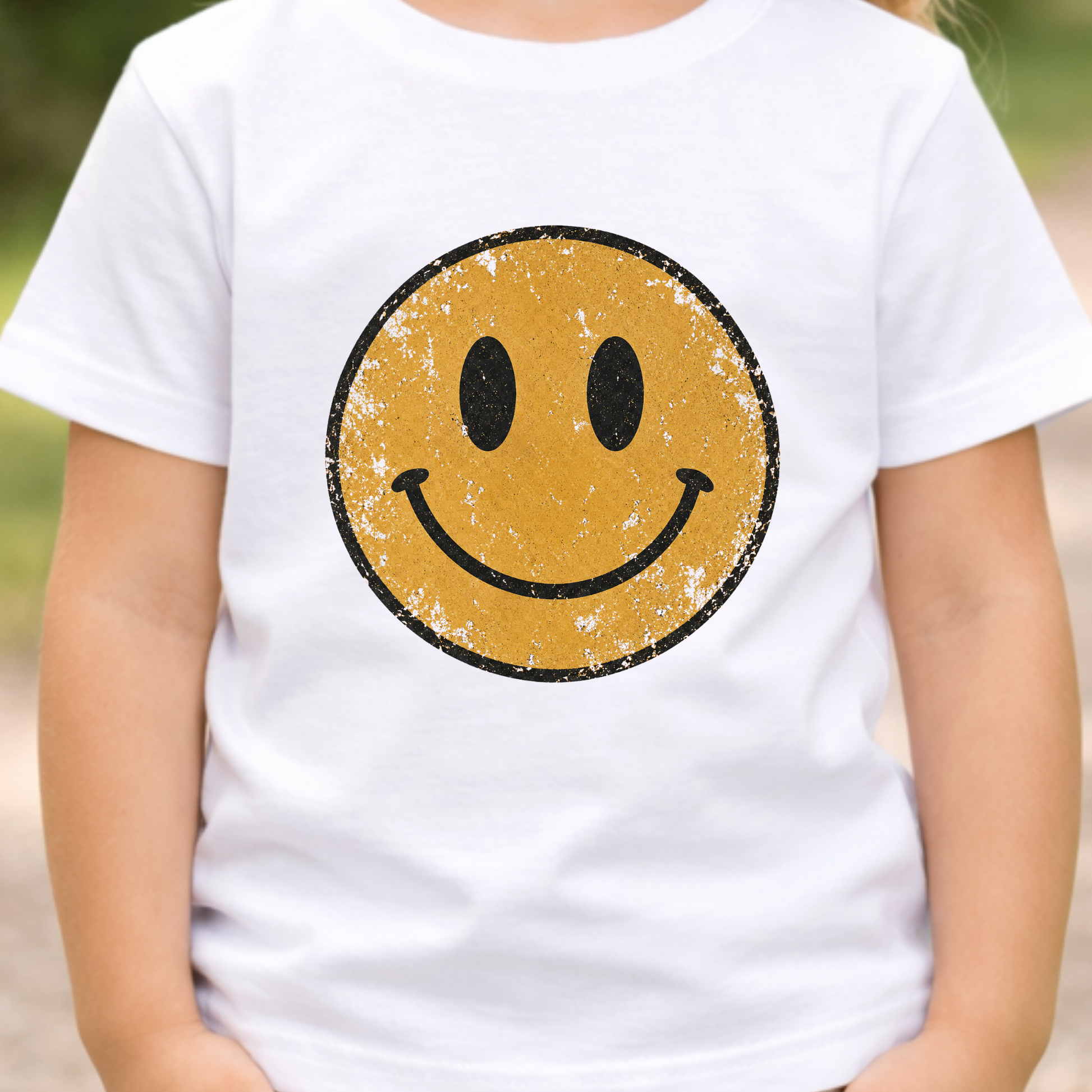 A child wearing a white short-sleeve T-shirt with a large distressed yellow smiley face graphic on the front, photographed outdoors with a blurred natural background.