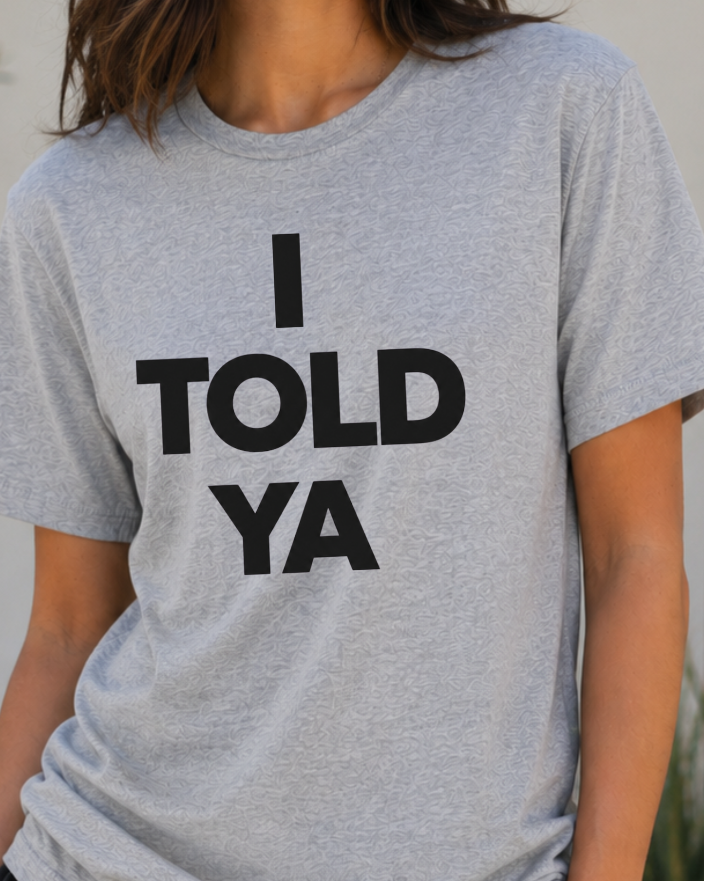 A close-up of a person wearing a heather gray short-sleeve t-shirt with the bold black text 'I TOLD YA' printed on the front.