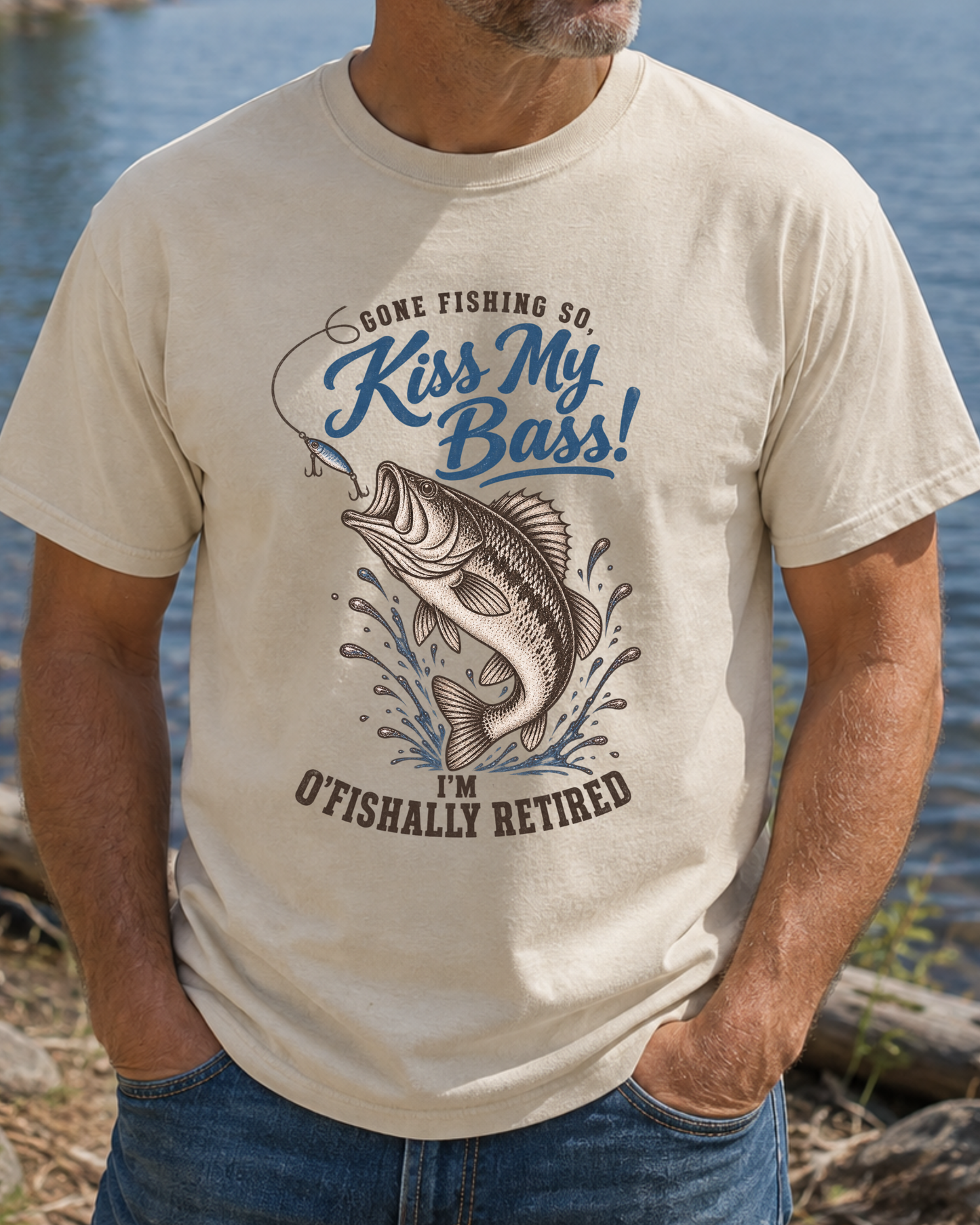 A man wearing a beige graphic t-shirt with a blue and brown fish design and the text 'Gone Fishing So, Kiss My Bass! I'm O'Fishally Retired', standing outdoors near water.
