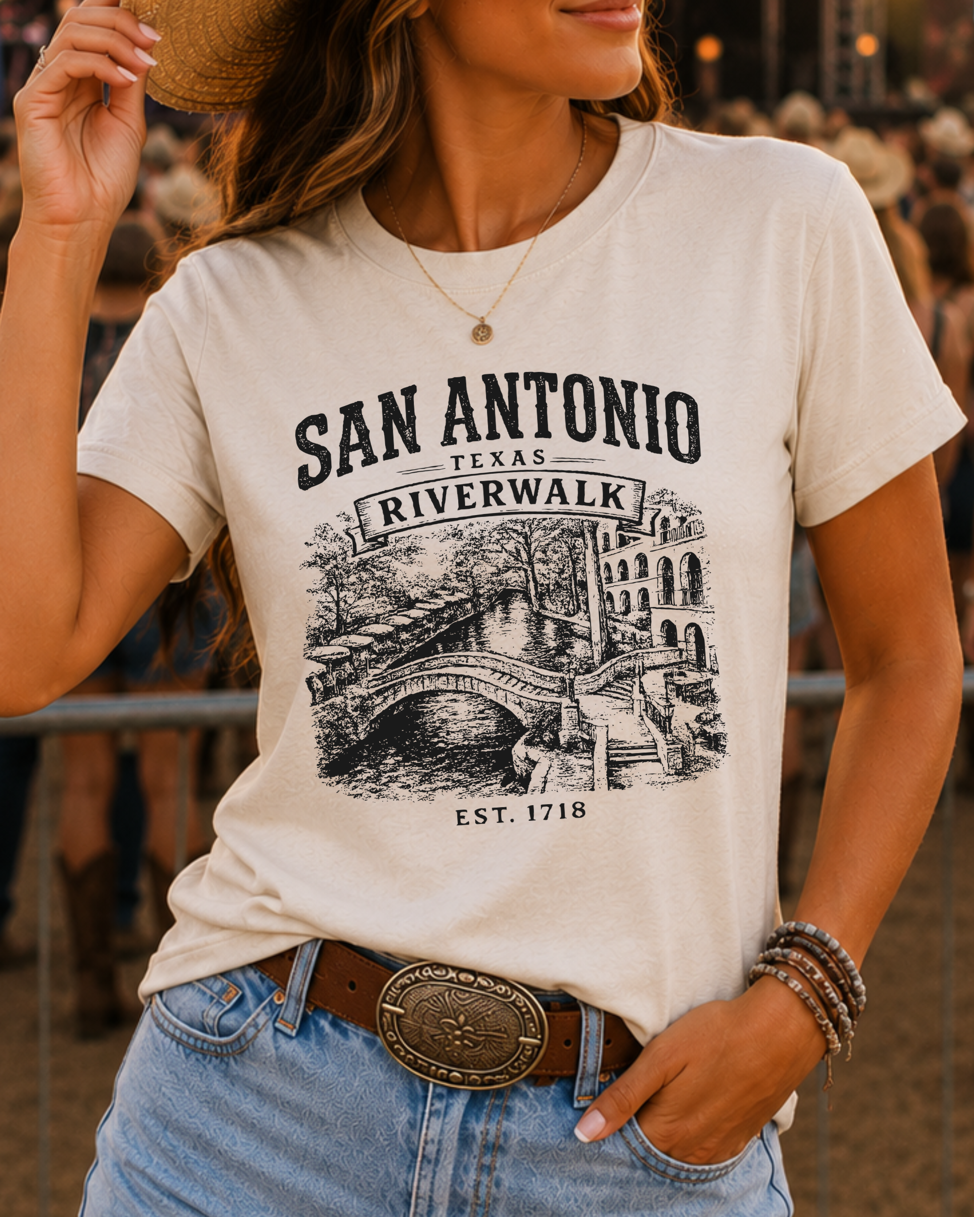 A woman wearing a cream-colored graphic t-shirt with a black print of the San Antonio Riverwalk, paired with light blue jeans and a brown belt.