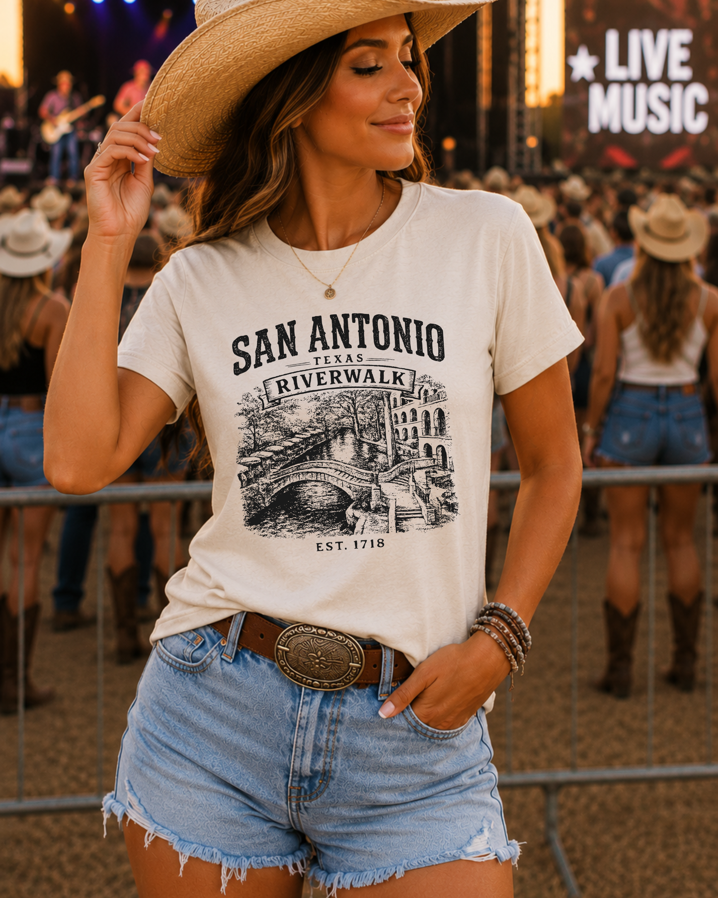 A woman wearing a cream-colored graphic t-shirt that reads 'SAN ANTONIO RIVERWALK EST. 1718' with a black illustration, paired with denim shorts and a cowboy hat, standing in a crowded outdoor concert setting.
