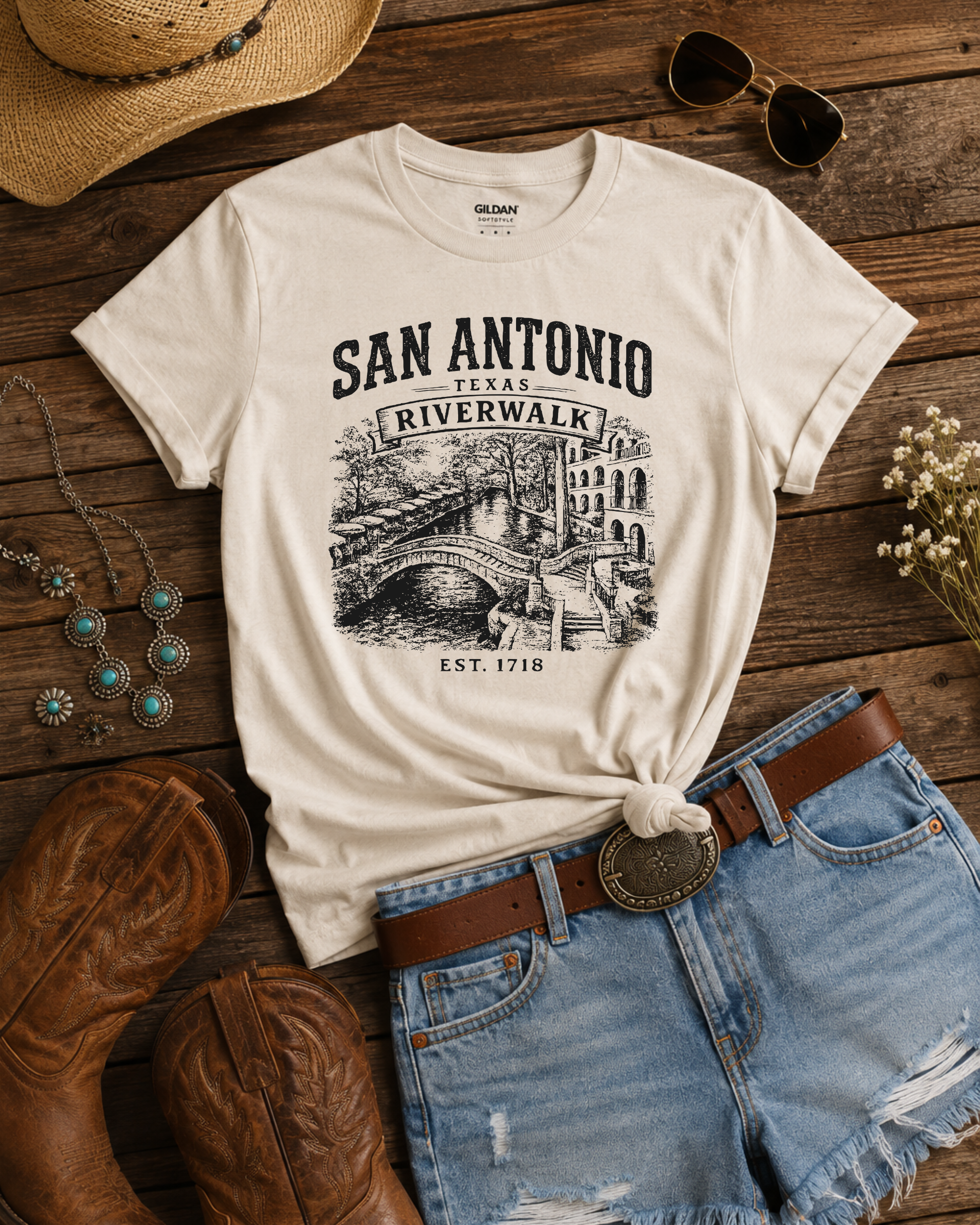 An off-white graphic t-shirt featuring a black print of the San Antonio Riverwalk, styled in a flat lay with denim shorts, a brown leather belt, cowboy boots, sunglasses, a straw hat, and turquoise jewelry on a rustic wooden background.