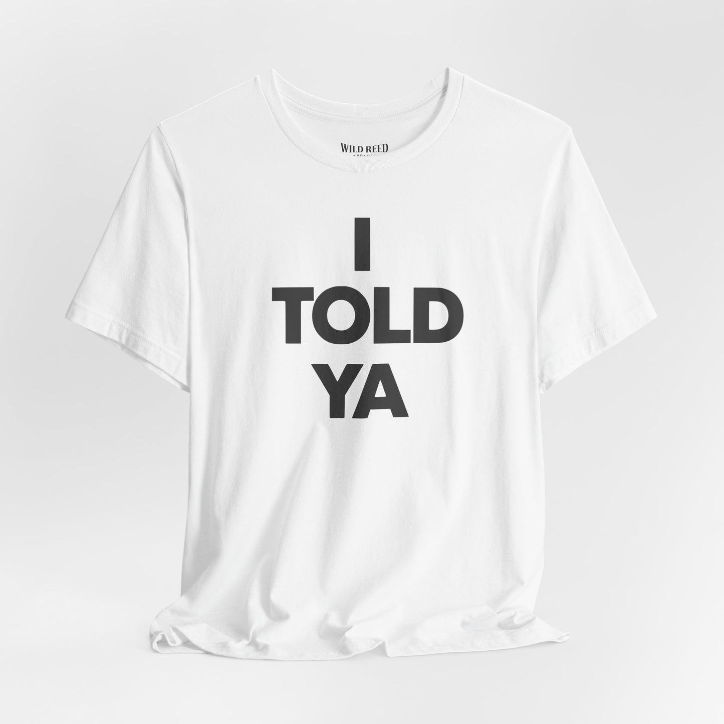 white t-shirt with the text 'I TOLD YA' printed in bold black letters, displayed on a clean white background. The inner collar shows the brand name 'WILD Reed'.